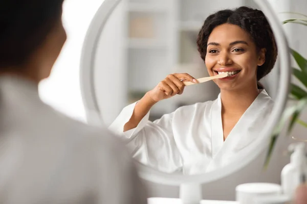 A woman wearing a bathrobe smiles while brushing her teeth.